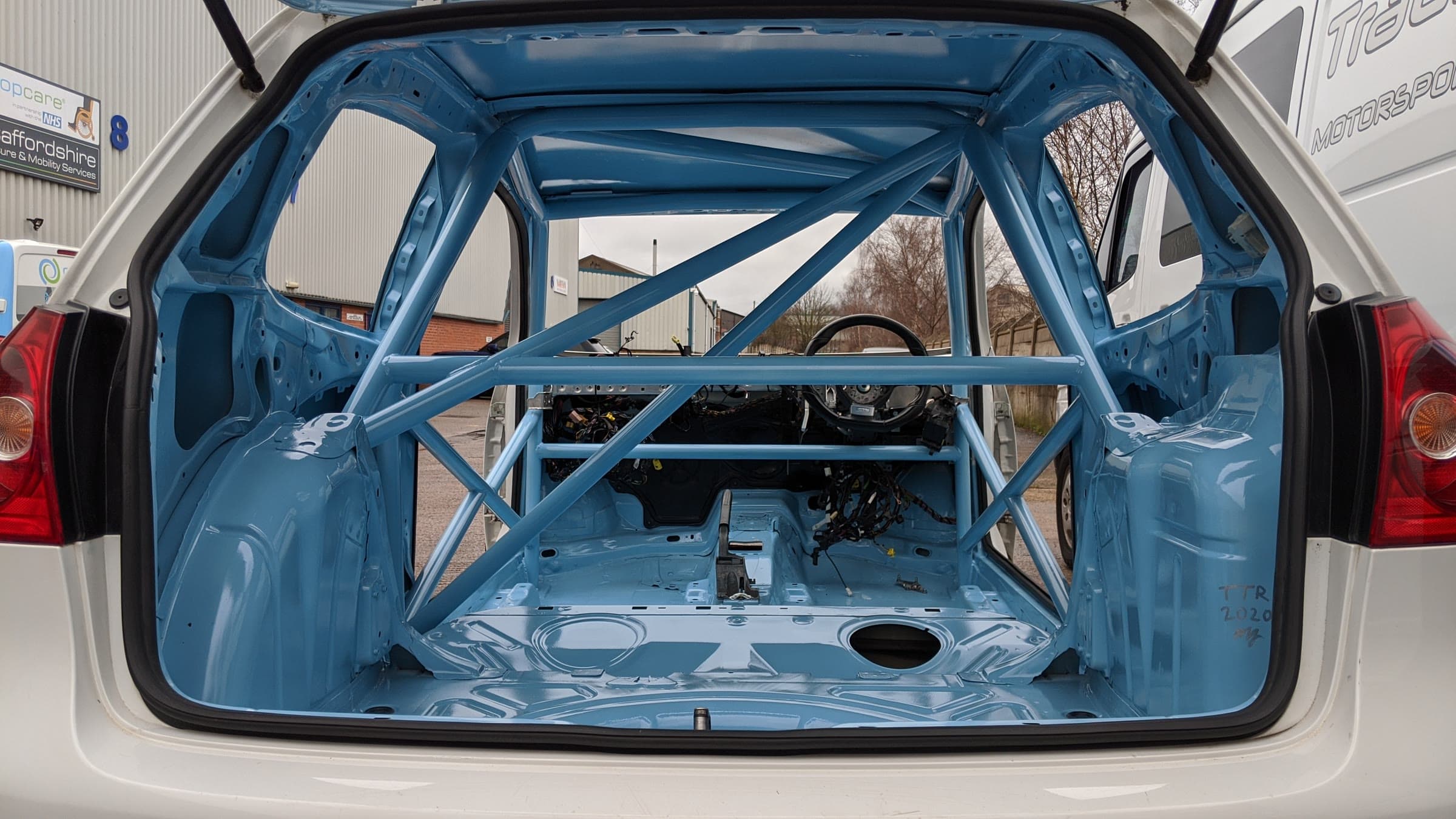 A white Golf build seen through the open boot: the full roll cage, painted blue, inside the stripped shell