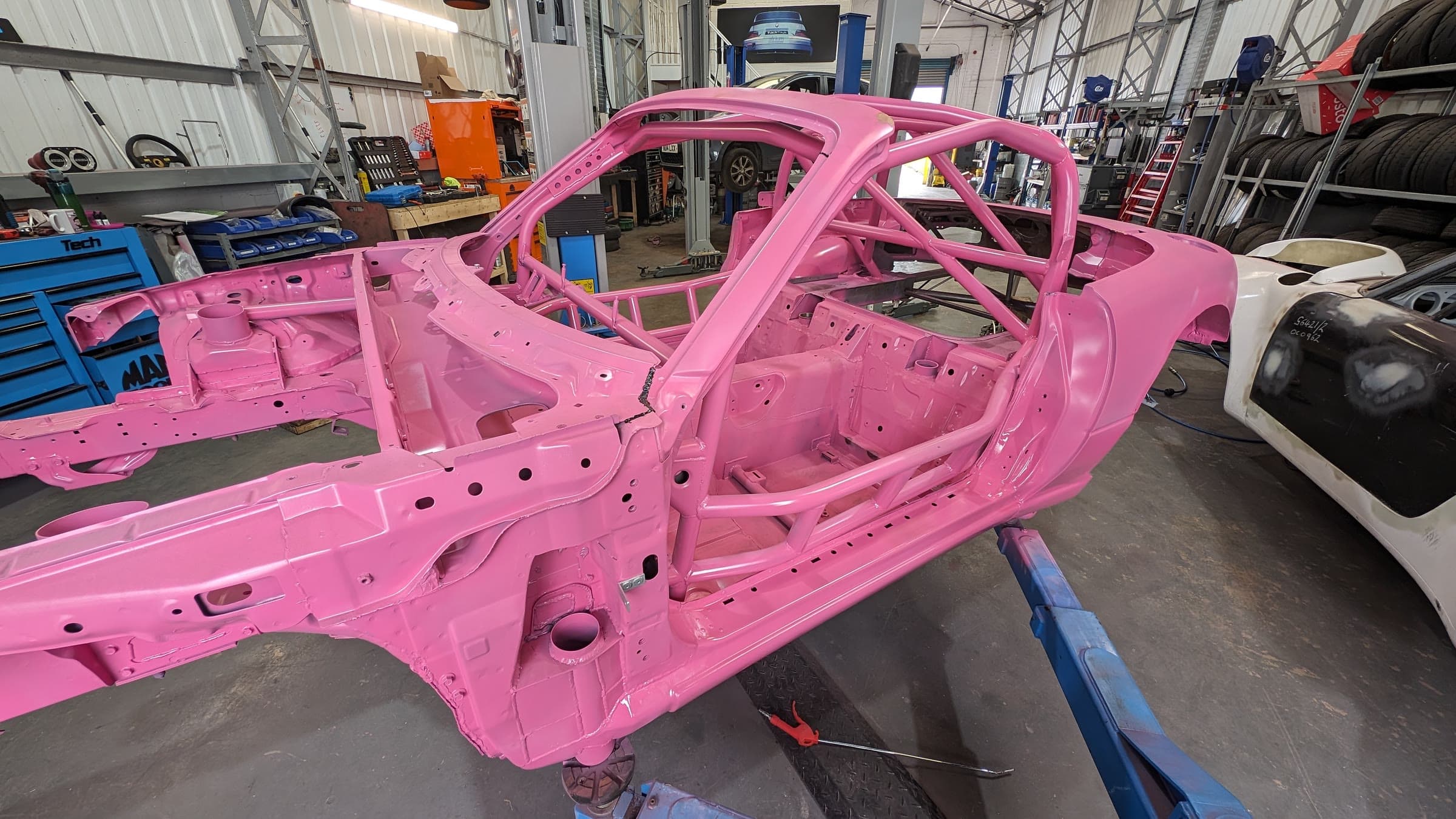 A pink Mazda MX5 race shell with a full roll cage, on a stand in the workshop